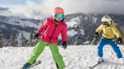 Zwei Kinder fahren auf einem verschneiten Berghang Ski und tragen bunte Winterjacken, Hosen und Helme.