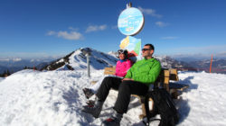 Zwei Personen in Skiausrüstung entspannen sich auf einer Bank in einer verschneiten Berglandschaft unter blauem Himmel.