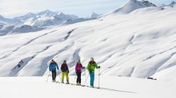 Vier Skitourengeher auf einem verschneiten Berghang mit schneebedeckten Gipfeln im Hintergrund.
