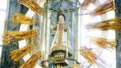 Die Statue eines gekrönten Kindes in prunkvollem Gewand, umgeben von goldenen Strahlen in einer Kirche, ist von Glas umgeben.