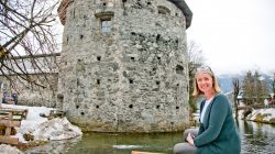 Frau sitzt auf einer Bank an einem verschneiten Bach mit einem runden Steinturm im Hintergrund.