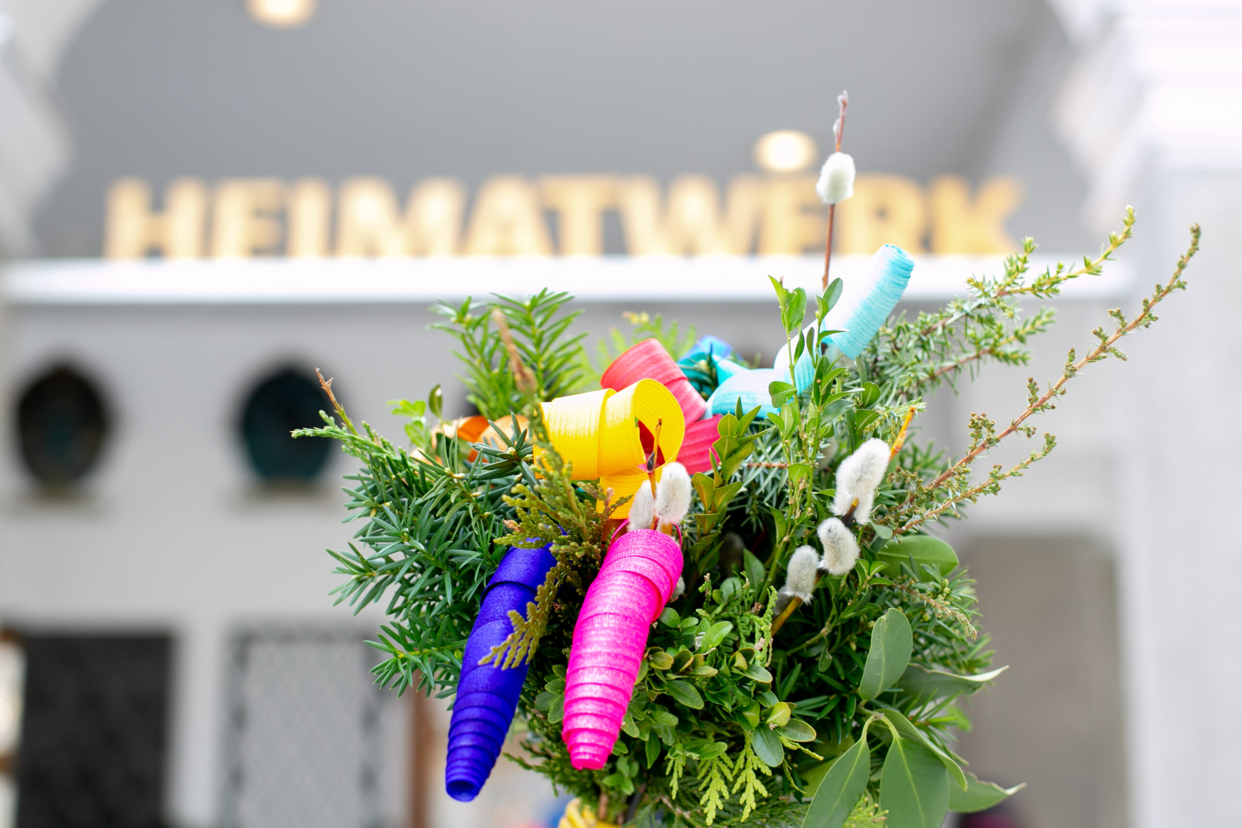 Ein bunter Blumenstrauß mit gekräuselten Papierdekorationen vor einem verschwommenen HEIMATWERK-Schild.
