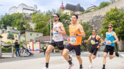 Vier männliche Läufer in Sportkleidung laufen auf einer Straße mit historischen Gebäuden und einem Schloss im Hintergrund.