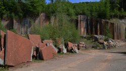 Große rötliche Steinplatten und -blöcke, die neben einer unbefestigten Straße gestapelt sind, mit Bäumen und einer felsigen Klippe im Hintergrund.