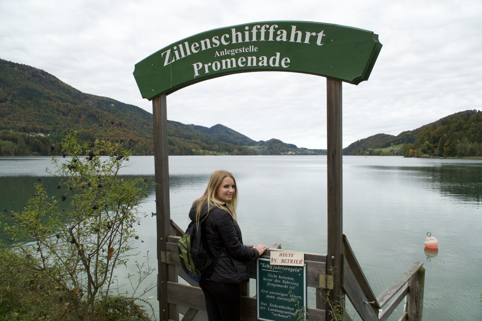 Eine Frau steht an einem See unter einem grünen Schild mit der Aufschrift Zillenschifffahrt Promenade mit Hügeln im Hintergrund.