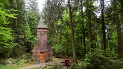 Kleine hölzerne Kapelle in einem üppigen grünen Wald, in dessen Nähe eine Person auf einer Bank sitzt.
