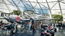 Ein glänzendes Red Bull-Flugzeug und ein Rennwagen sind in einem modernen Glasgebäude mit gewölbten Decken ausgestellt.