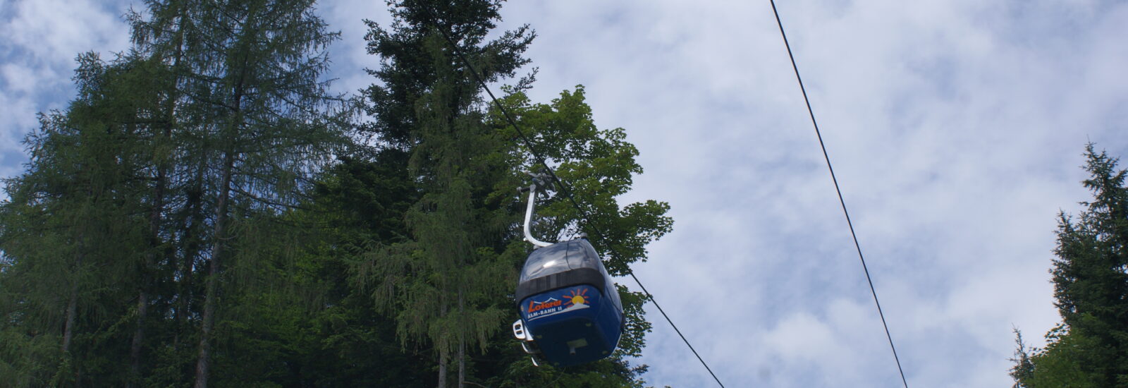 Eine blaue Seilbahn fährt vor einem teilweise bewölkten Himmel über hohen grünen Bäumen hinweg.