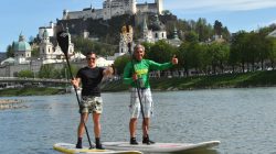 Zwei Männer paddeln auf einem Fluss und zeigen die Daumen nach oben, mit einer Stadt und einem Schloss auf einem Hügel im Hintergrund.