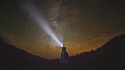Eine Person mit einer Stirnlampe steht unter einem sternenklaren Nachthimmel, der Lichtstrahl ist nach oben gerichtet.