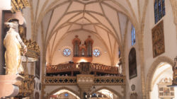 Das Innere einer historischen Kirche mit gewölbten Decken, Orgel, Statuen und einem sitzenden Publikum mit Blick nach vorne.