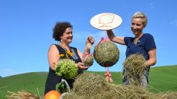 Zwei Frauen im Freien mit Heudekoration an einem Tisch mit Blumen, Gemüse und einem Schild unter blauem Himmel.