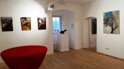 Galerieraum mit Holzböden, Wandkunst, einem roten runden Tisch und einem Fenster mit einer kleinen Skulptur.