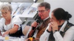 Drei ältere Erwachsene in traditioneller österreichischer Tracht sitzen an einem Tisch und unterhalten sich; ein Mann spricht in ein Mikrofon.