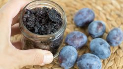 Eine Hand hält ein Glas mit Pflaumenmus neben mehreren frischen Pflaumen auf einer gewebten Fläche.