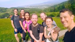 Sieben lächelnde Freunde posieren für ein Selfie auf einer malerischen Wanderung mit Hügeln und einer Stadt im Hintergrund.