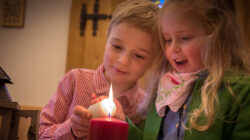 Zwei kleine Kinder zünden gemeinsam eine rote Kerze an, lächelnd, in einem gemütlichen Raum.
