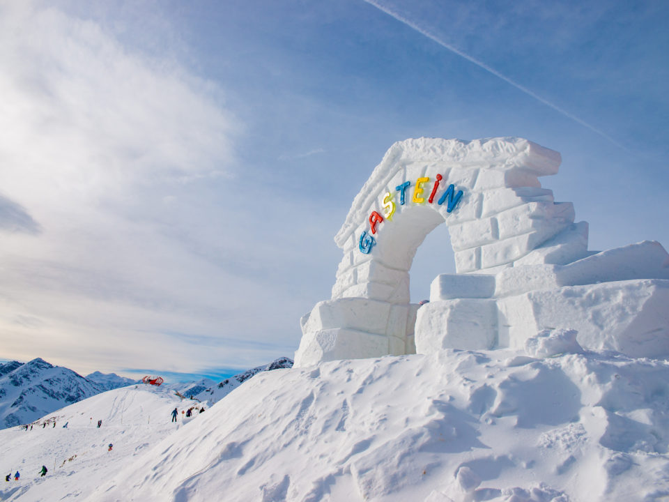 Ein großer Schneebogen mit bunten GASTEIN-Buchstaben steht auf einem verschneiten Berg mit Menschen und Gipfeln im Hintergrund.
