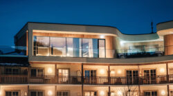 Modernes Hotelgebäude mit großen Fenstern und warmen Lichtern, die nachts vor einem klaren, dunkelblauen Himmel leuchten.