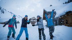 Vier Personen in Winterkleidung lächeln und spielen im Schnee und halten ein Matte Mach Hills-Schild neben verschneiten Hütten.