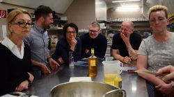 Sechs Erwachsene versammeln sich um einen Küchentisch mit Getränken, einer Flasche und Essen und scheinen sich zu unterhalten.