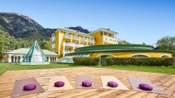 Vier Yogamatten mit lilafarbenen Polstern auf einer Holzplattform vor einem gelben Hotel in einer Bergregion.