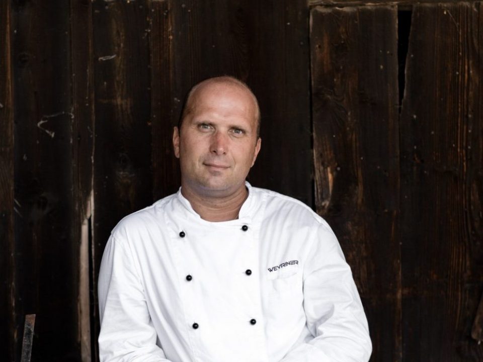 Ein Koch im weißen Kittel sitzt an einer rustikalen Holzwand und schaut ruhig in die Kamera.