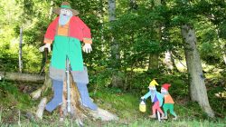 Bunte Holzfiguren eines bärtigen Mannes und zweier Zwerge auf einer Waldlichtung.