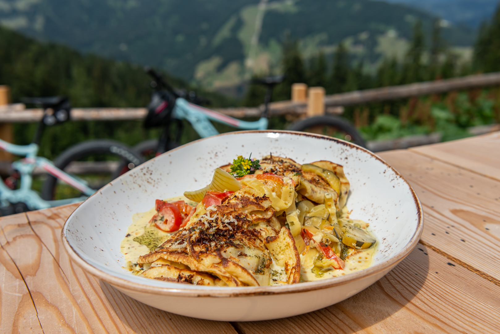 Ein Teller mit herzhaften Crêpes mit Gemüse und Soße auf einem Tisch im Freien, Berge und Fahrräder im Hintergrund.