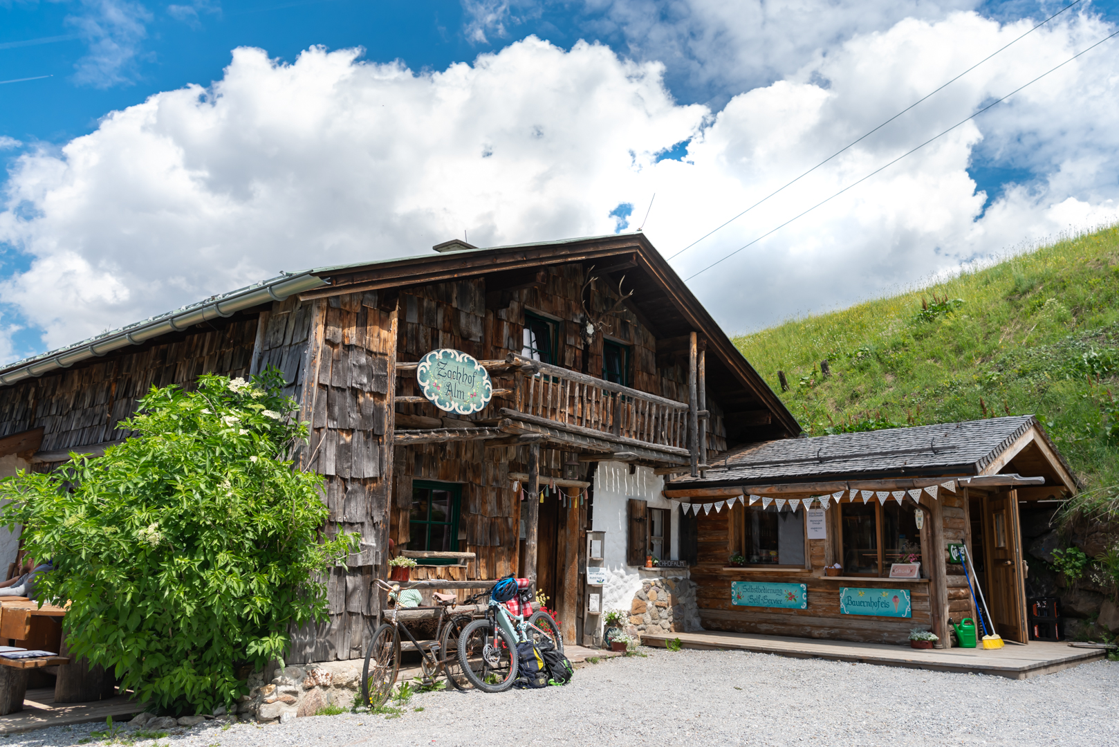Eine rustikale Holzhütte mit einer kleinen Veranda, Fahrräder im Freien und ein grasbewachsener Hügel unter einem teilweise bewölkten Himmel.