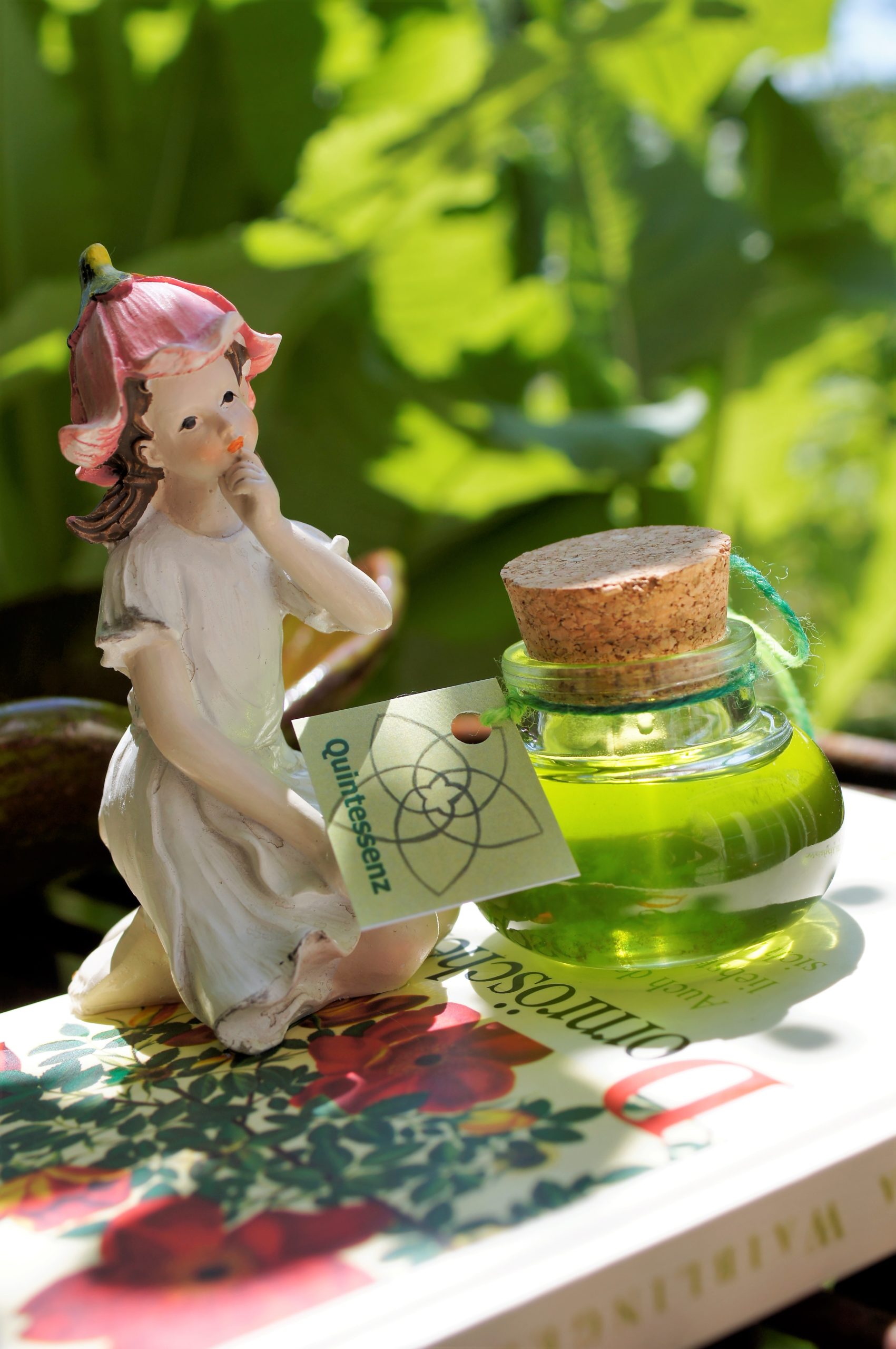 Eine blumengekrönte Figur sitzt neben einer kleinen grünen Zaubertrankflasche mit einem Korken und einem Anhänger auf einem Buch im Freien.