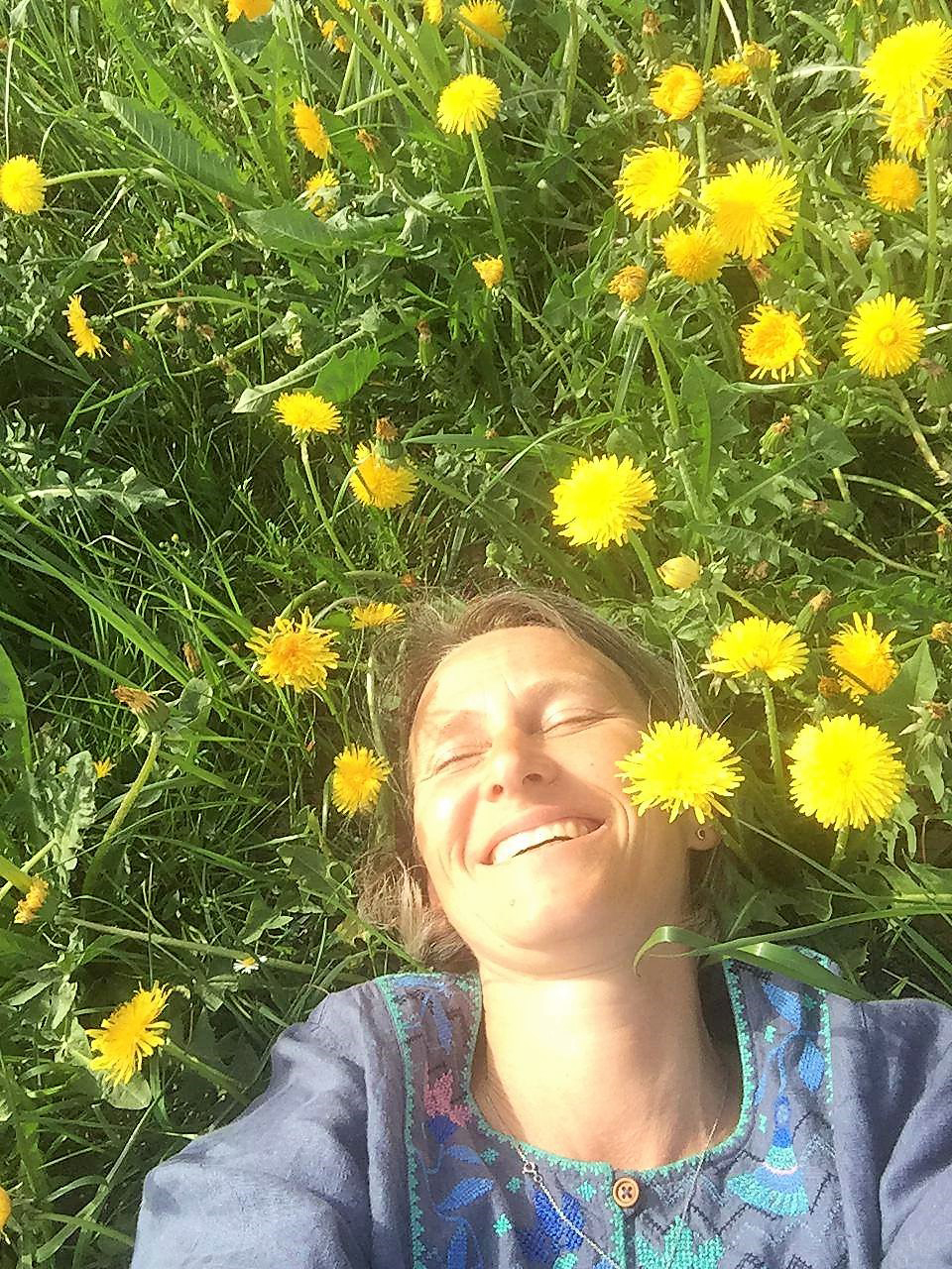 Eine lächelnde Person liegt an einem sonnigen Tag im Gras, umgeben von gelbem Löwenzahn.