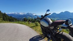 Ein Postlam-Motorrad, geparkt auf einer Bergstraße mit malerischen Hügeln und blauem Himmel im Hintergrund.