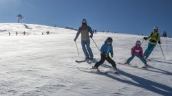 Eine Gruppe, darunter auch Kinder, fährt in einem Familienskigebiet bei strahlend blauem Himmel einen verschneiten Hang hinunter.