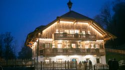 Ein warm beleuchtetes Holzchalet, Historisches Apartmenthaus Plainstöckl, mit Balkonen und Menschen am Eingang in der Abenddämmerung.