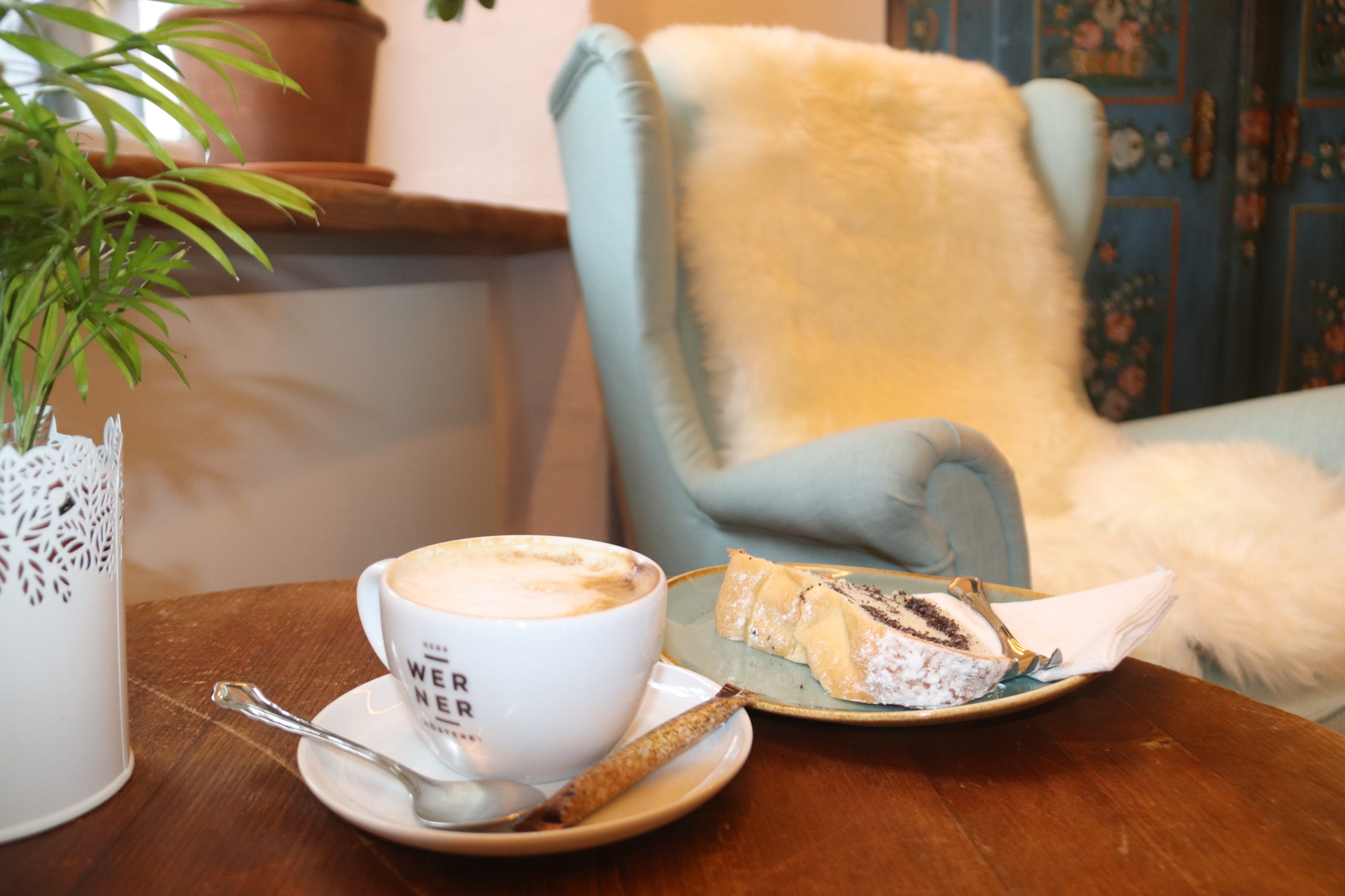 Cappuccino und Kuchen auf einem Tisch neben einem gemütlichen Sessel - Die Tauglerei ist ein Zentrum für Ayurveda und mehr.