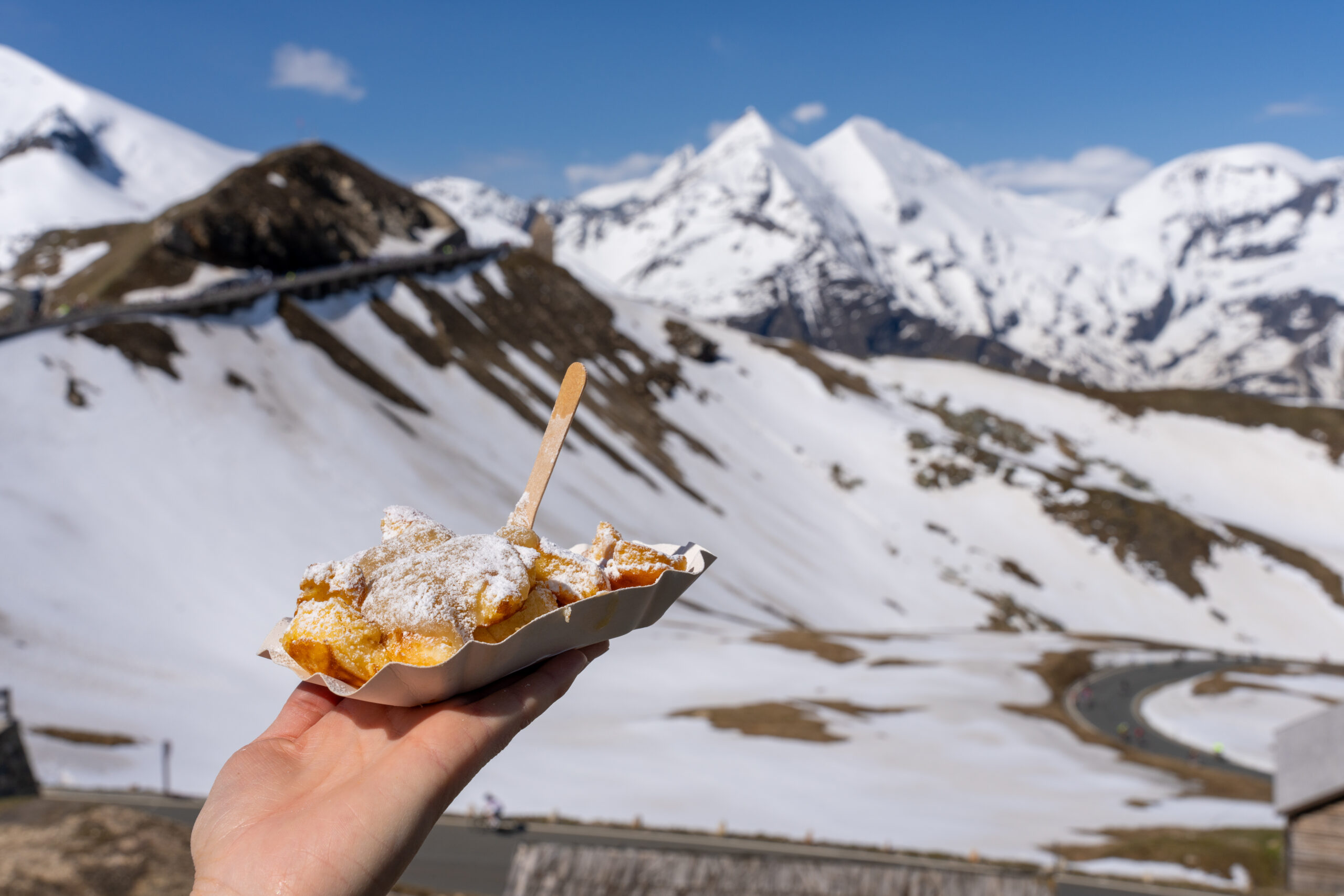 Eine Hand hält gepudertes Gebäck am Glocknerkönig, verschneite Berge leuchten an einem sonnigen Tag.