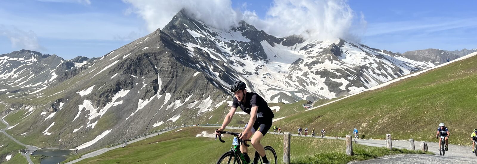 Ein Radfahrer fährt auf einer Bergstraße am Glocknerkönig bergauf, mit schneebedeckten Gipfeln und Wolken im Hintergrund.