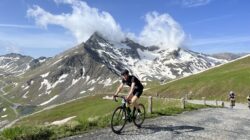Ein Radfahrer fährt auf einer Bergstraße am Glocknerkönig bergauf, mit schneebedeckten Gipfeln und Wolken im Hintergrund.
