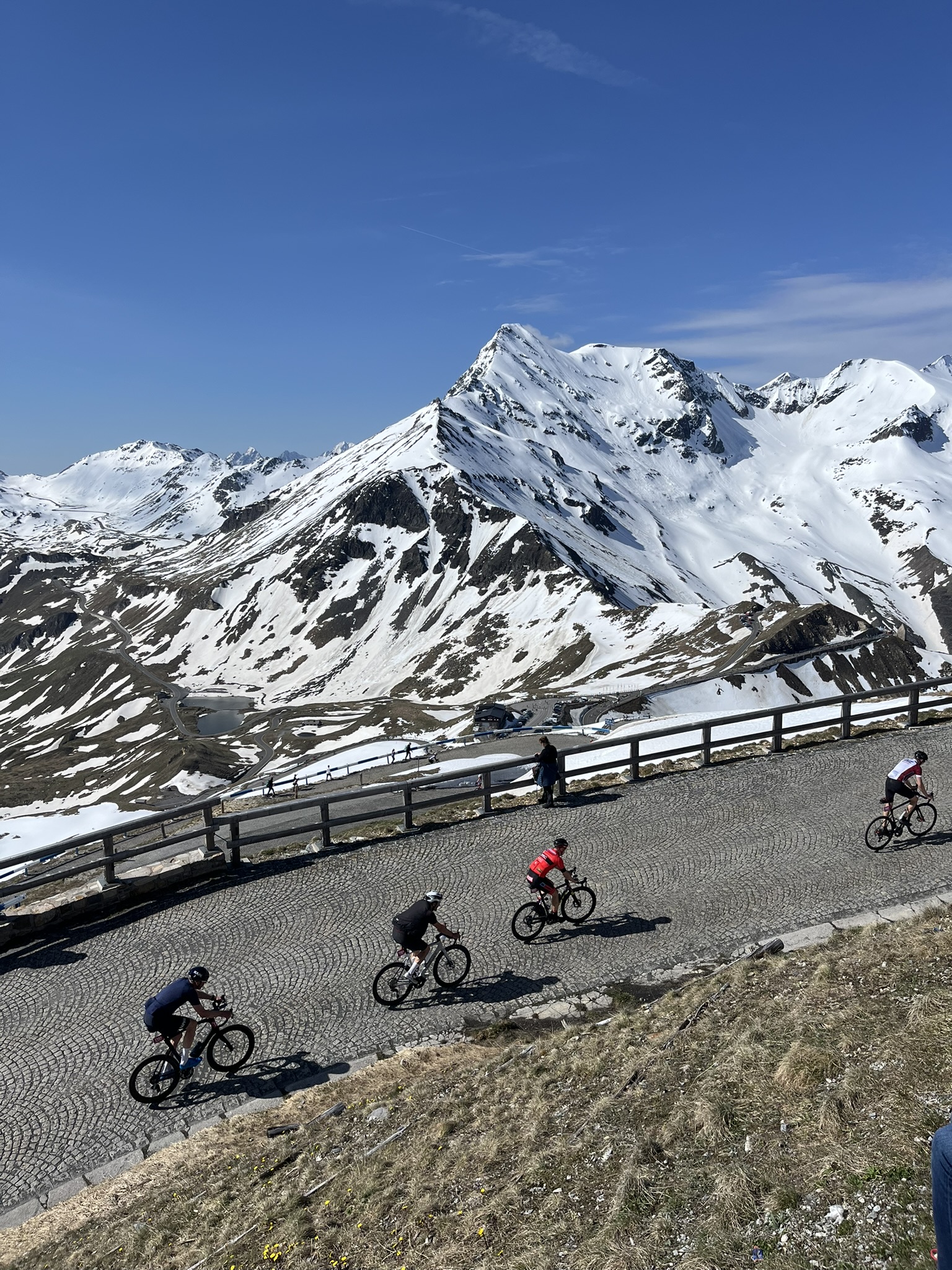 Radfahrer fahren beim malerischen Glocknerk&ouml;nig eine kurvenreiche Bergstra&szlig;e hinauf, dahinter Schneegipfel und blauer Himmel.