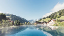 Infinity-Pool mit Blick auf die Berge, umgeben von Häusern und Grünanlagen unter einem klaren blauen Himmel.