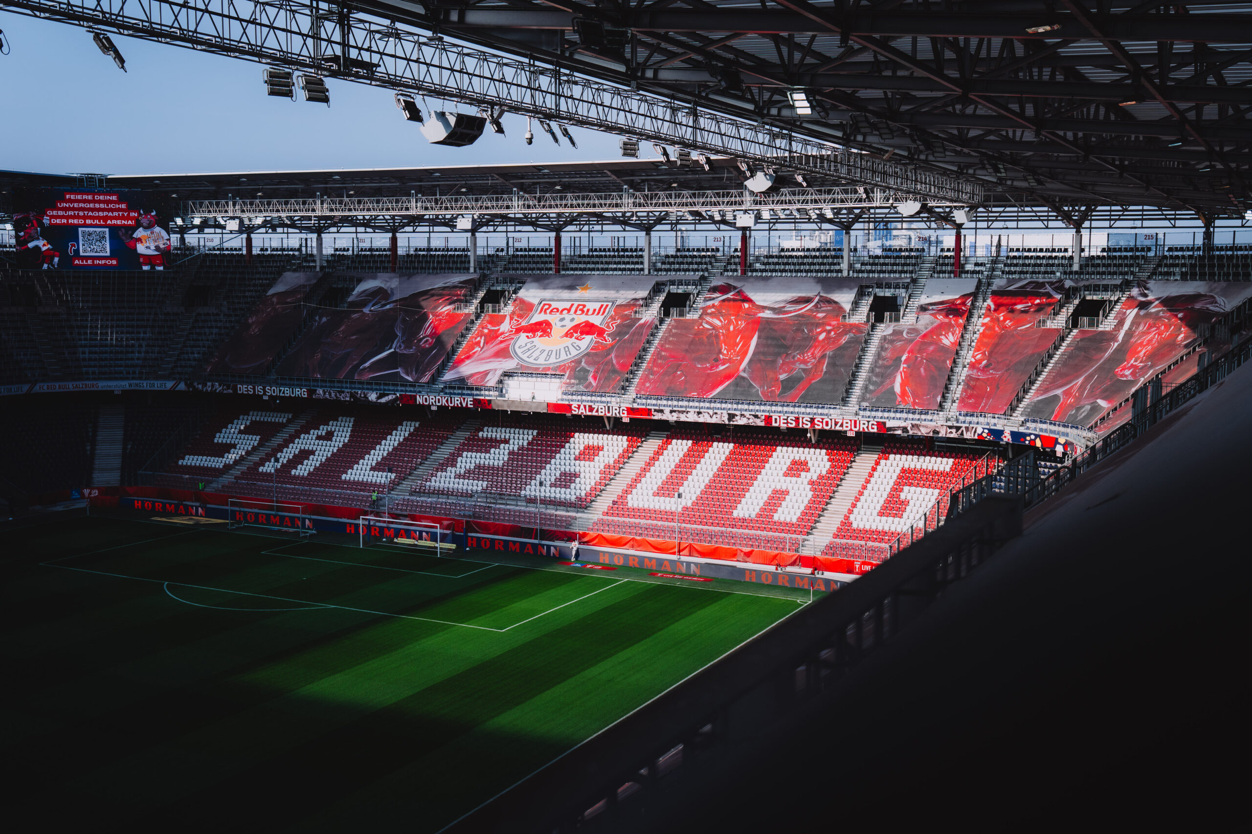 Die leeren Sitzplätze im Fußballstadion bilden das Wort „SALZBURG“, darüber hängen Banner von Red Bull Salzburg, und das Sonnenlicht strömt herein.