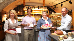 Vier Personen in einer Bäckerei, lächelnd und mit Geschenken, einem Kuchen und Brot in der Hand, stehen an einem Tresen mit Lebensmitteln.