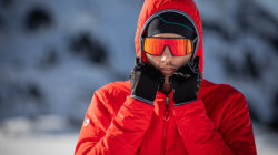 Person in roter Jacke, Handschuhen und reflektierender Sonnenbrille in verschneiter Umgebung.
