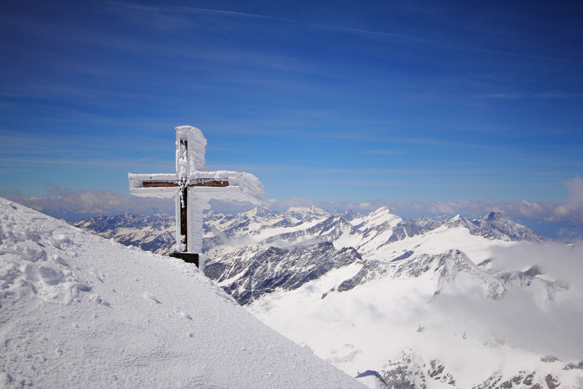 Ein schneebedecktes Kreuz steht auf einem Berggipfel mit verschneiten Bergen und blauem Himmel im Hintergrund.