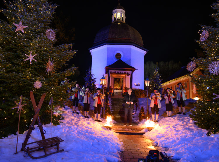 Eine Band spielt nachts vor einer Kapelle, umgeben von beleuchteten Weihnachtsbäumen, Schnee und festlicher Dekoration.