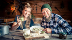 Ein lächelnder Mann und eine lächelnde Frau stellen an einem rustikalen Holztisch im Haus selbstgemachte Speisen und Kräuter aus.