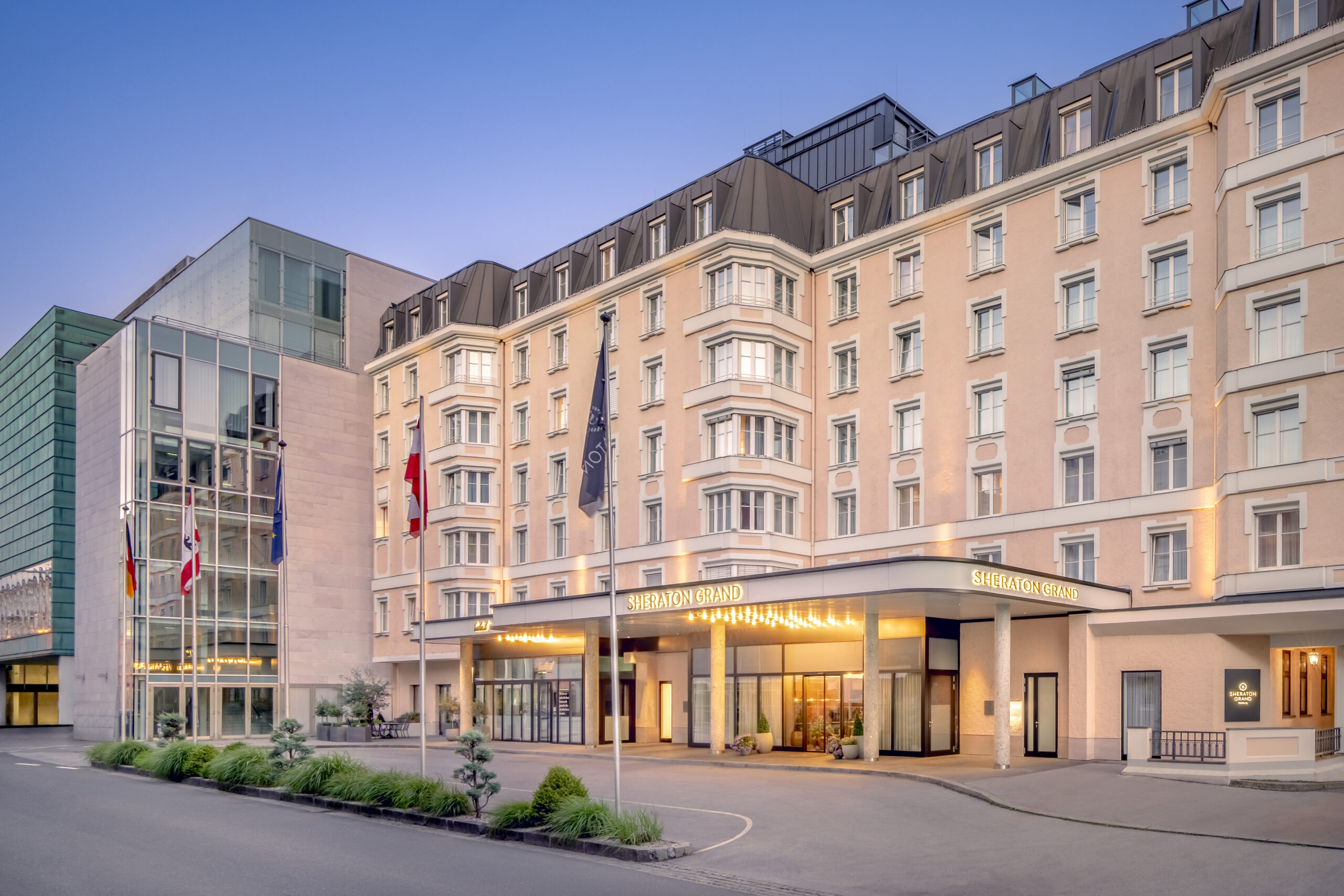 Sheraton Grand Salzburg von außen mit beleuchtetem Eingang, Fahnen und modernem Glasgebäude in der Abenddämmerung.