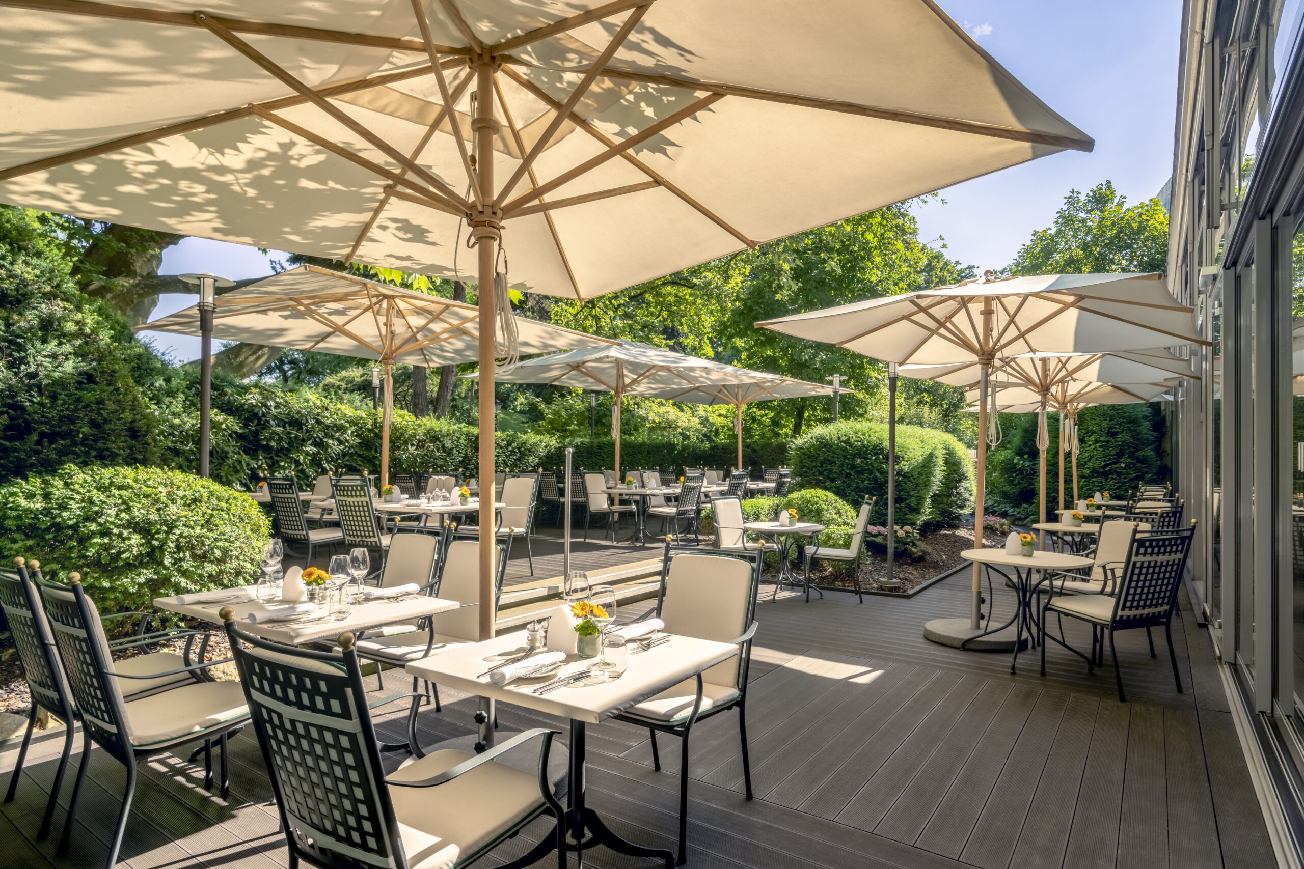 Sonnige Restaurantterrasse im Sheraton Grand Salzburg mit Tischen, Stühlen, Sonnenschirmen und üppigem Grün.