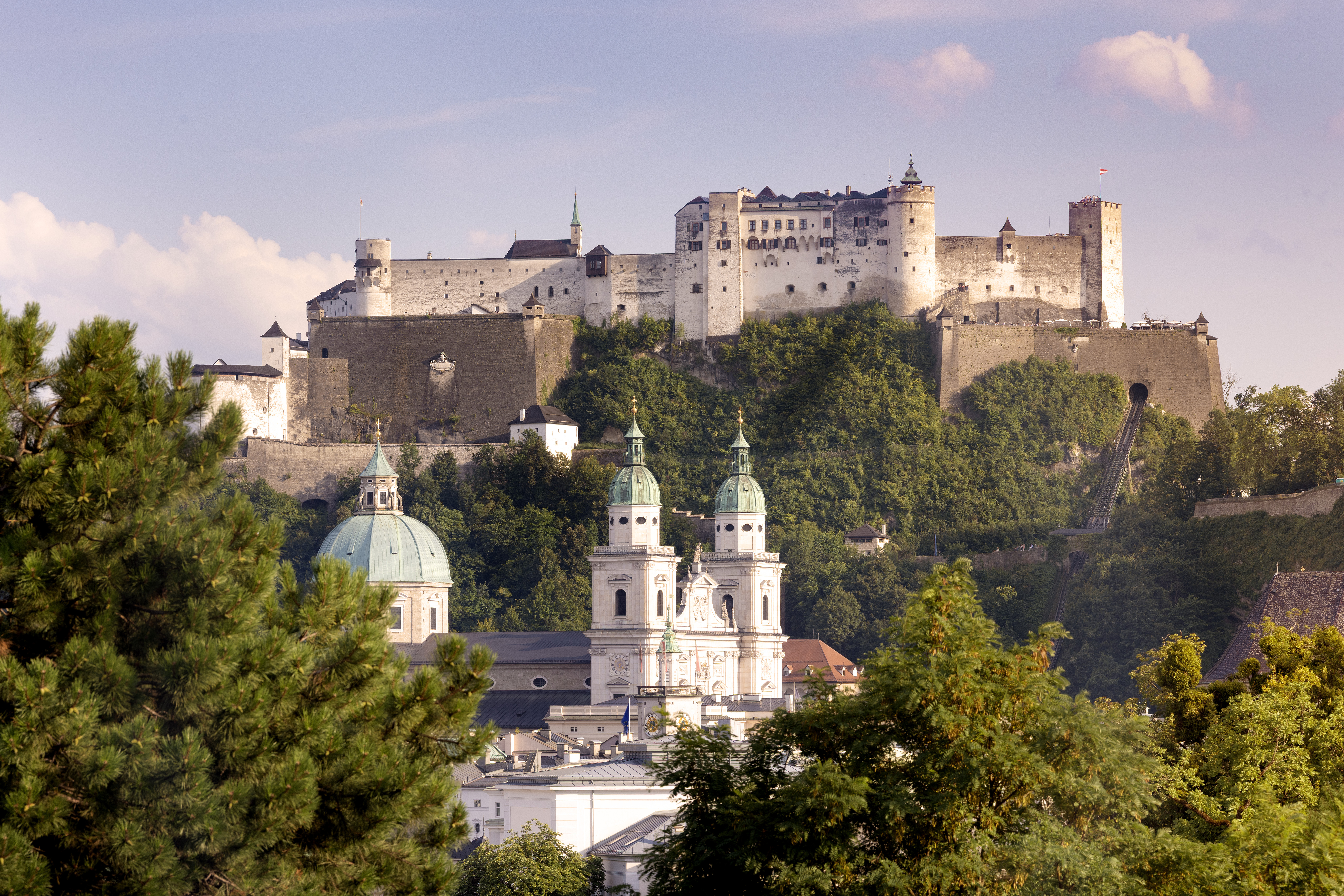 Eine große Festung auf einem Hügel überragt die Stadt und das Sheraton Grand Salzburg inmitten von Kuppeln und üppigen grünen Bäumen.